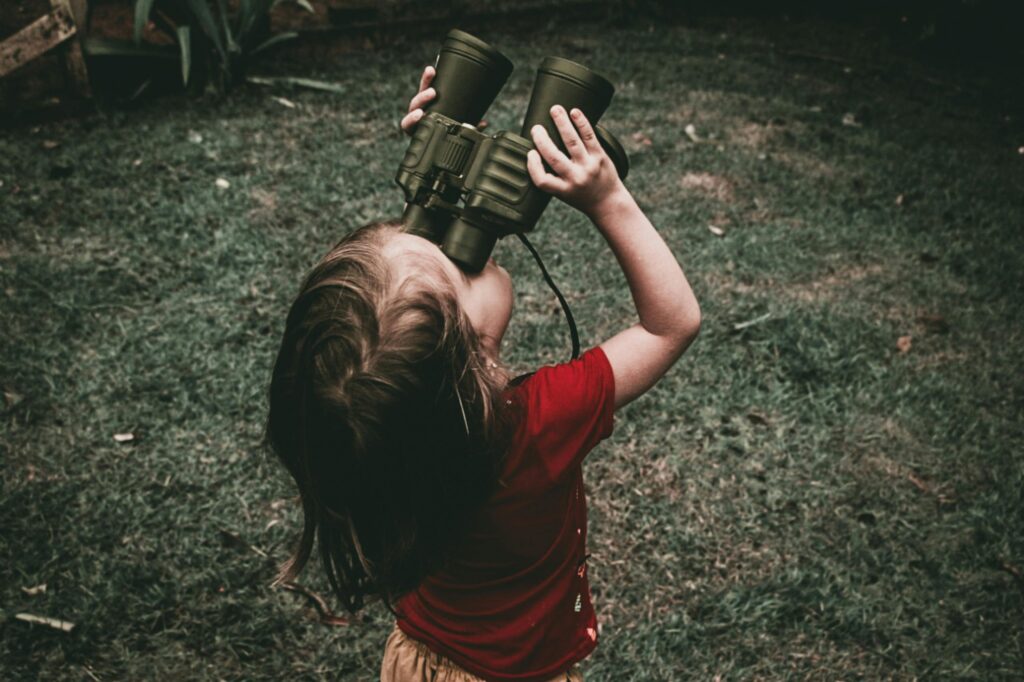 A young girl in a red shirt using binoculars to explore nature outdoors.